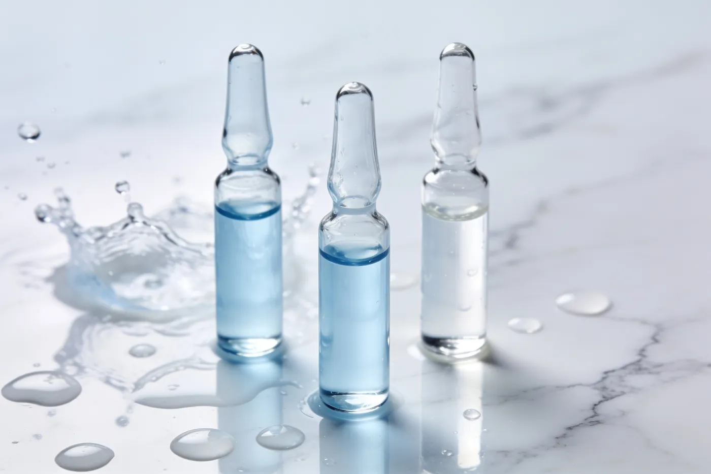 Three small glass ampoules of clear and pale-blue hyaluronic acid solution standing on white marble